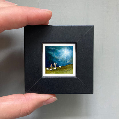 A person's hand holding a small square picture frame containing a painting of a pastoral scene with sheep and a starry sky.