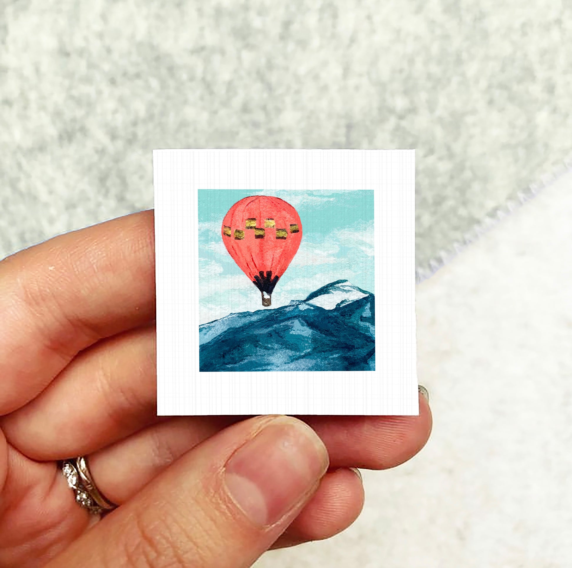 A hand holding a small square picture depicting a hot air balloon floating above a mountain landscape.