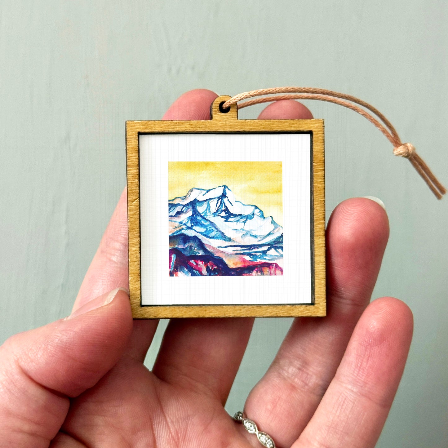 A hand holding a small wooden frame containing a watercolor painting of a mountain landscape.