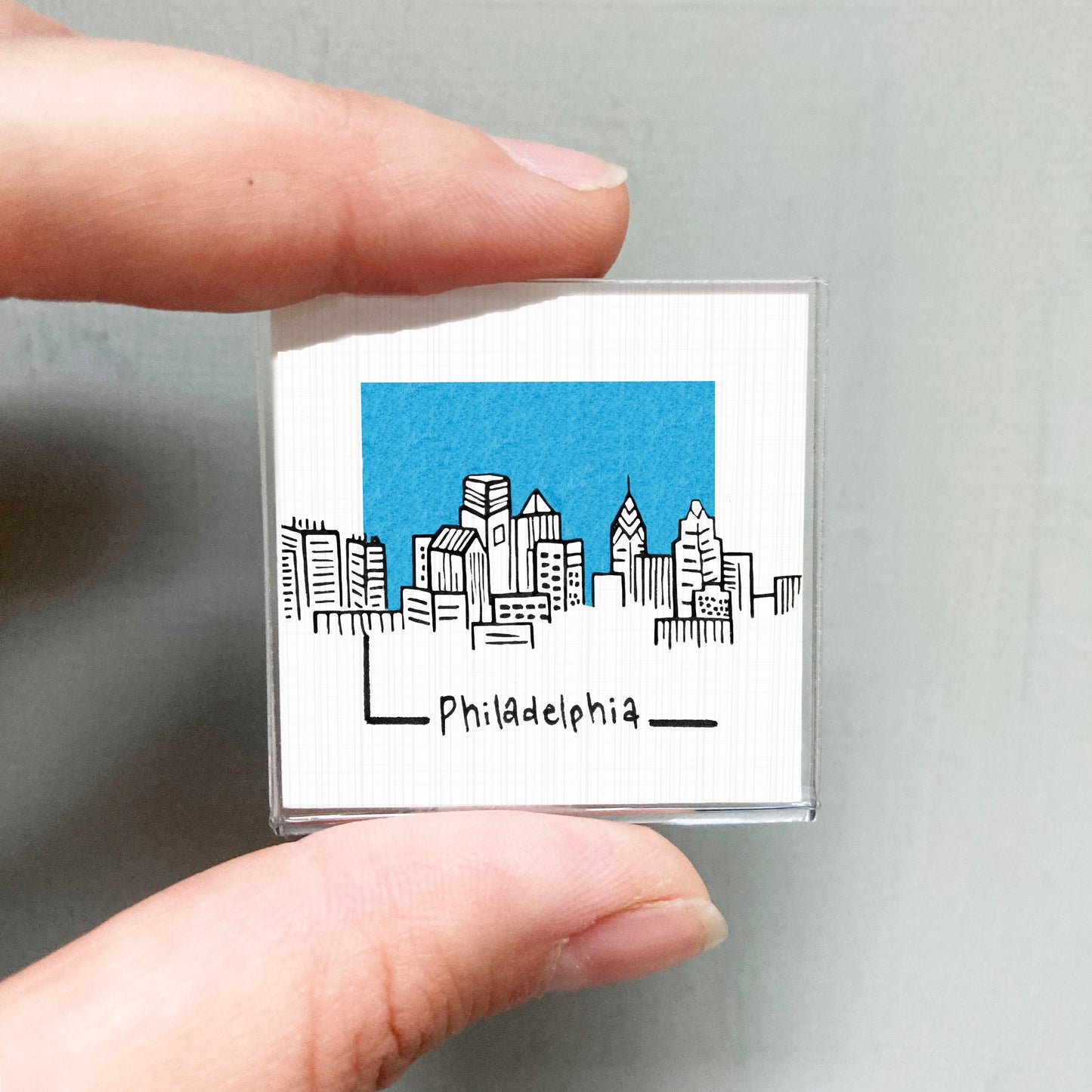 A hand holding a small square magnet with a drawing of a city skyline and the word "PhiladELPHIA" written below it.