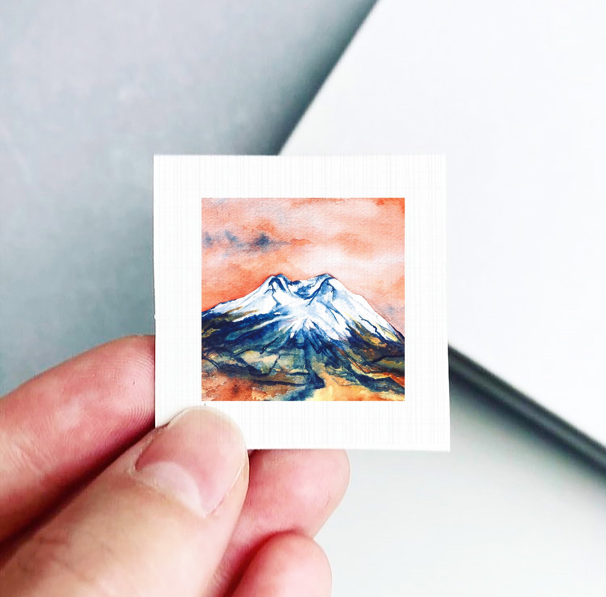 A hand is holding a small square artwork depicting a mountain landscape with a vibrant orange and blue sky.