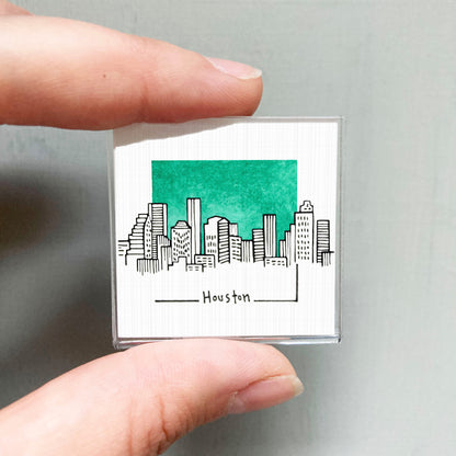 A hand holding a small square with a drawing of a city skyline and the word "Houston" written below it.