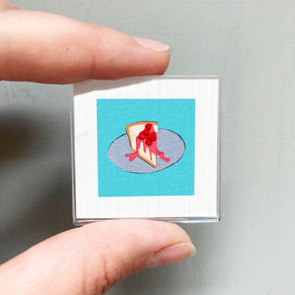 A person's hand holding a small square with a picture of a slice of cheesecake on a blue background.