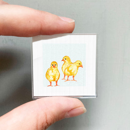 A person's hand is holding a small square with a painting of three yellow chicks on it.