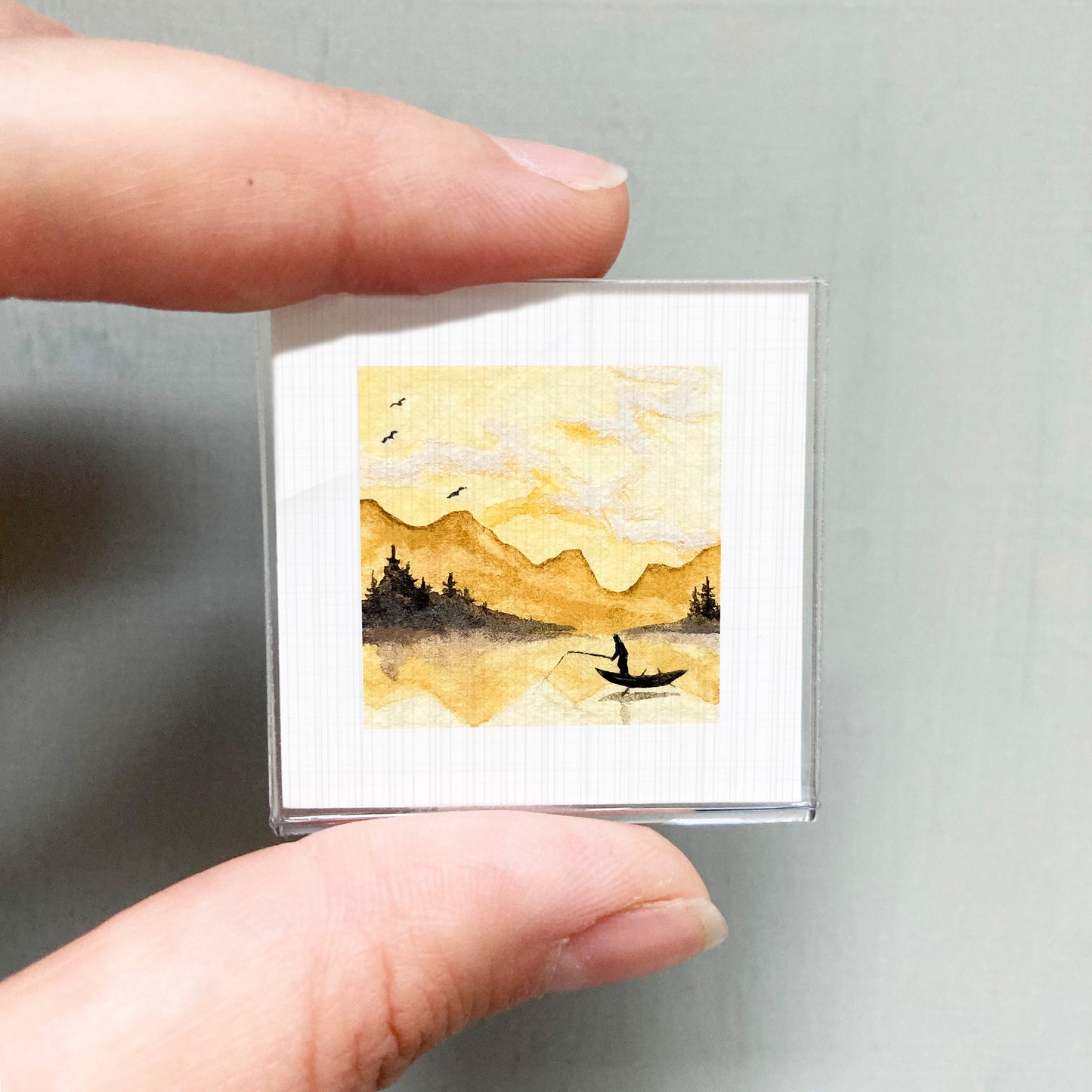 A person's hand is holding a small square picture depicting a serene landscape with a person fishing in a lake.