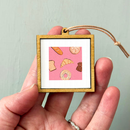 A person's hand holding a small wooden frame containing an illustration of various pastries, including bread, croissants, and donuts.