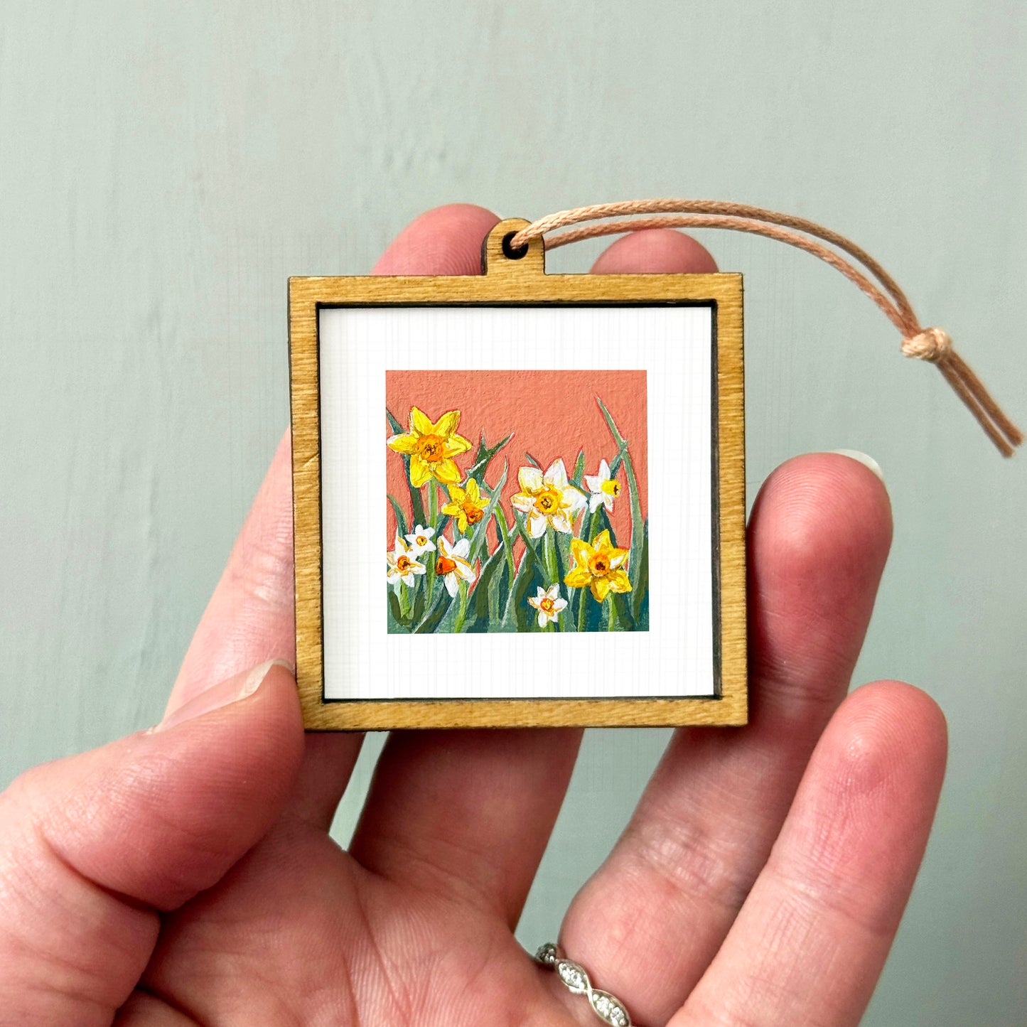 A hand holding a small wooden frame containing a painting of yellow daffodils.