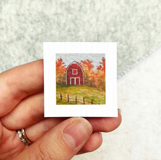 A hand holding a small white square with a painting of a red barn and autumn trees in the background.