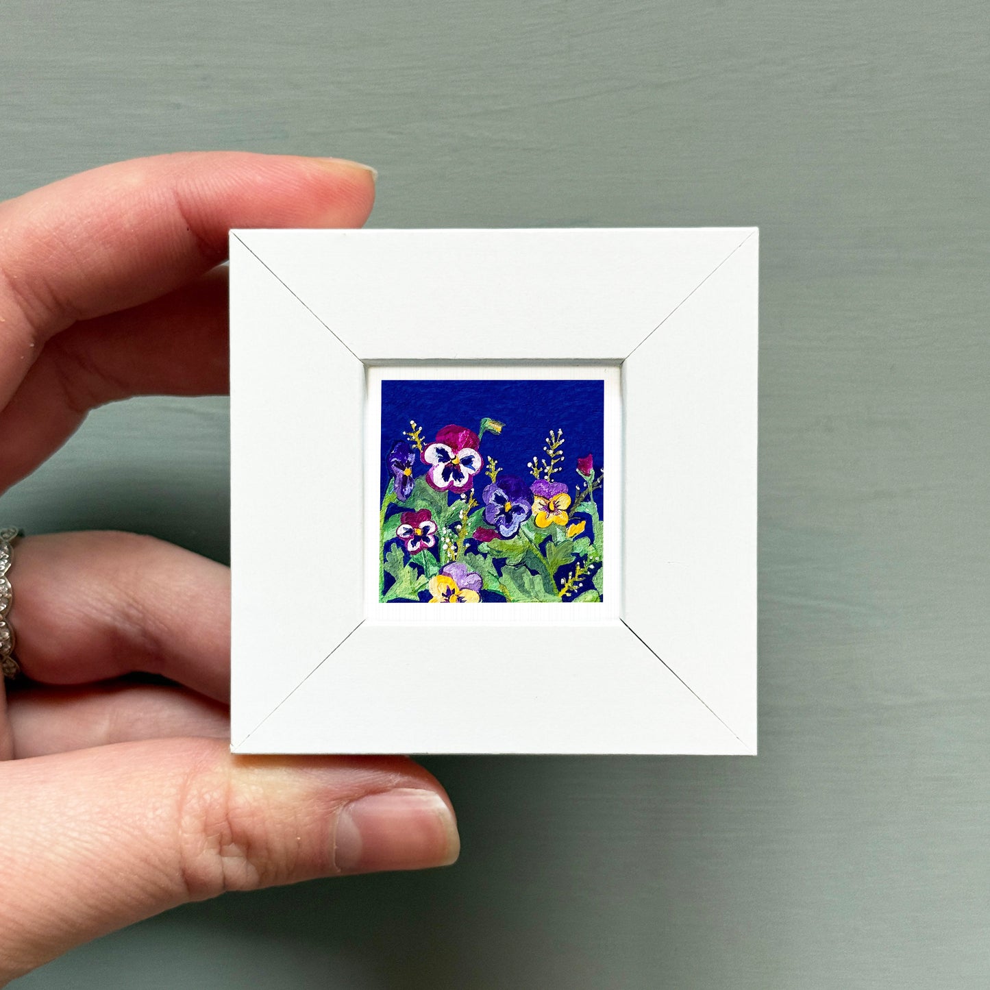 A hand is holding a small white frame containing a vibrant painting of colorful flowers against a dark blue background.