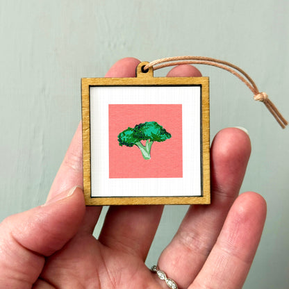 A hand holding a small wooden frame containing a framed image of a green broccoli floret.