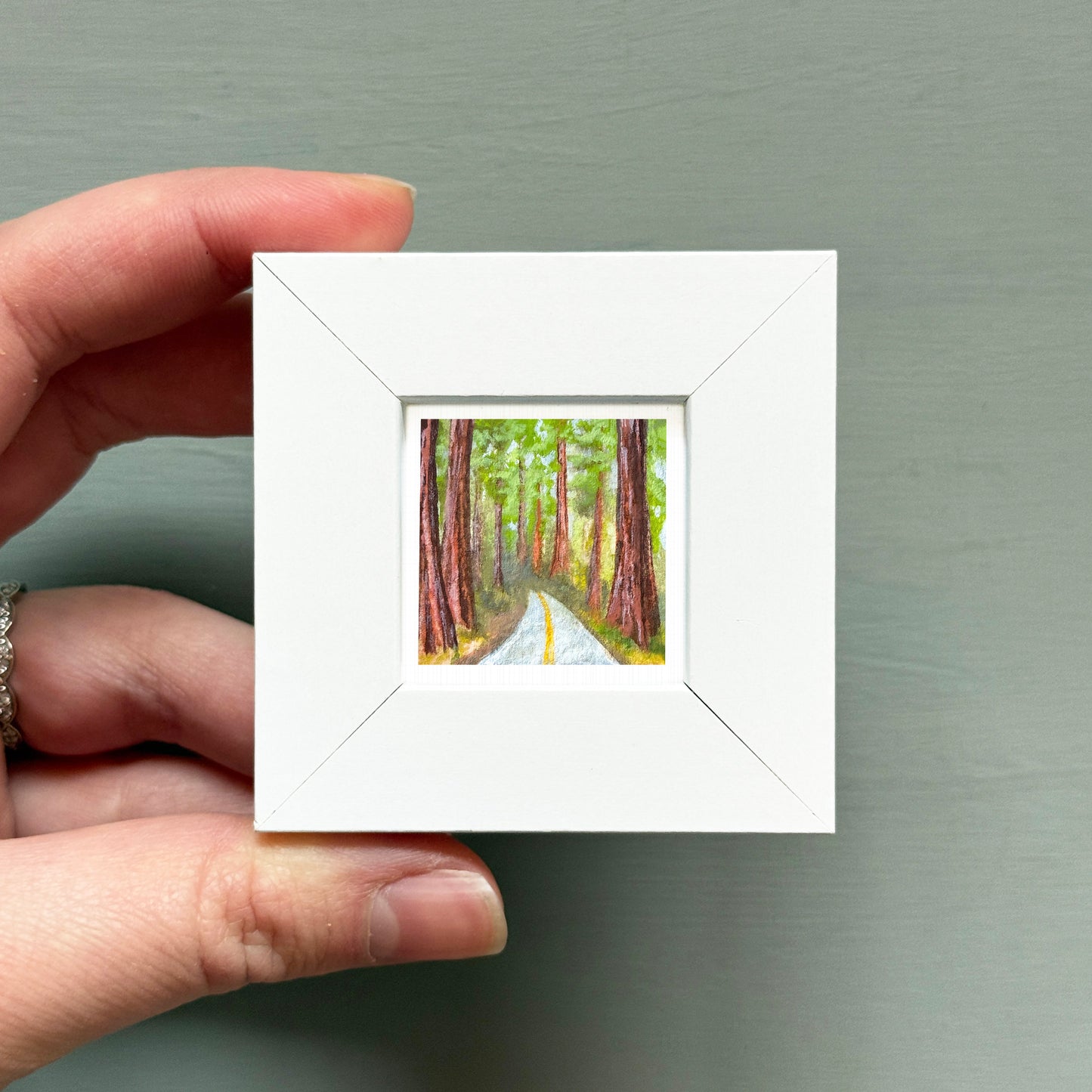 A hand is holding a small white square frame containing a painting of a forest road with tall trees.