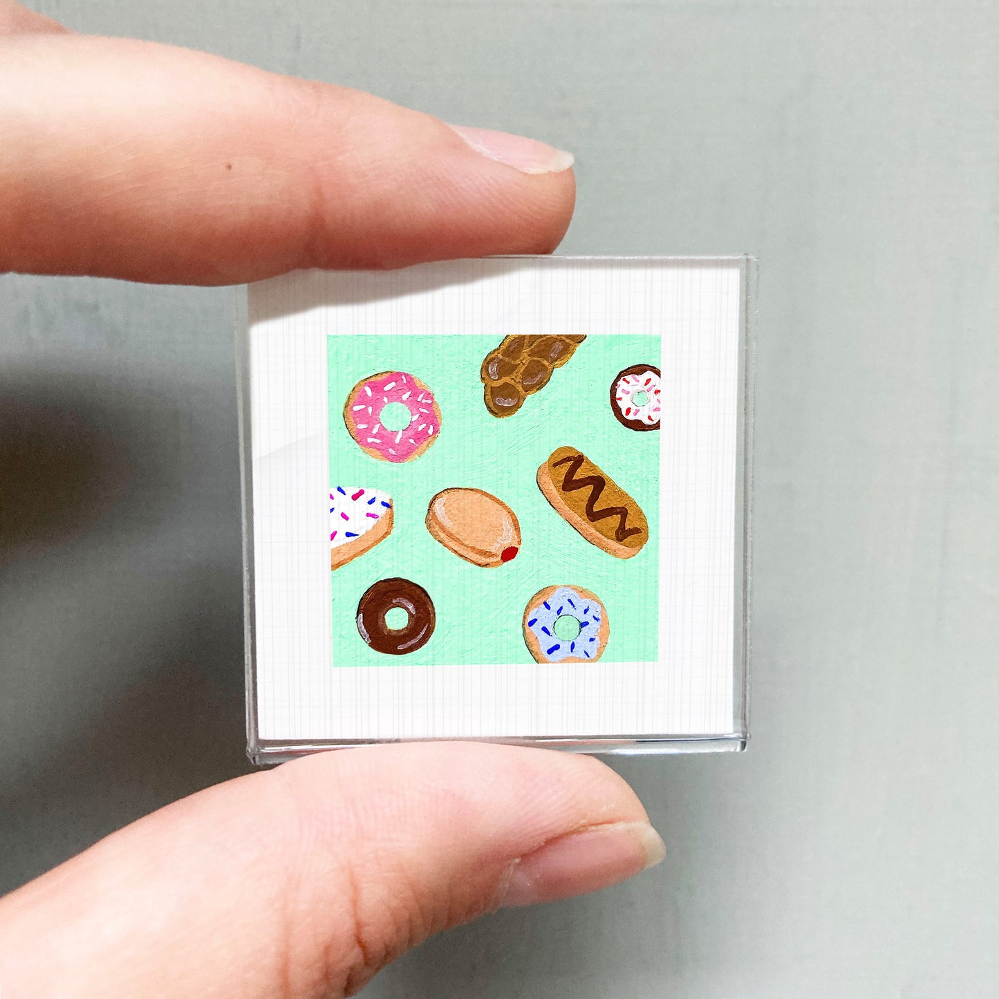 A person's hand is holding a small square with a vibrant illustration of various donuts on it.