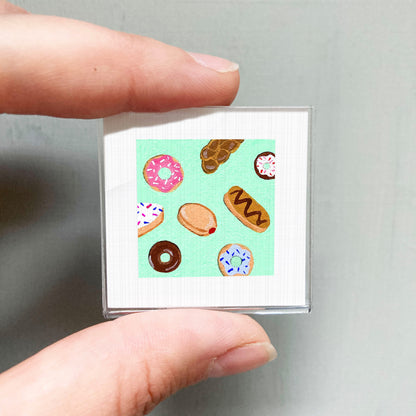 A person's hand is holding a small square with a vibrant illustration of various donuts on it.