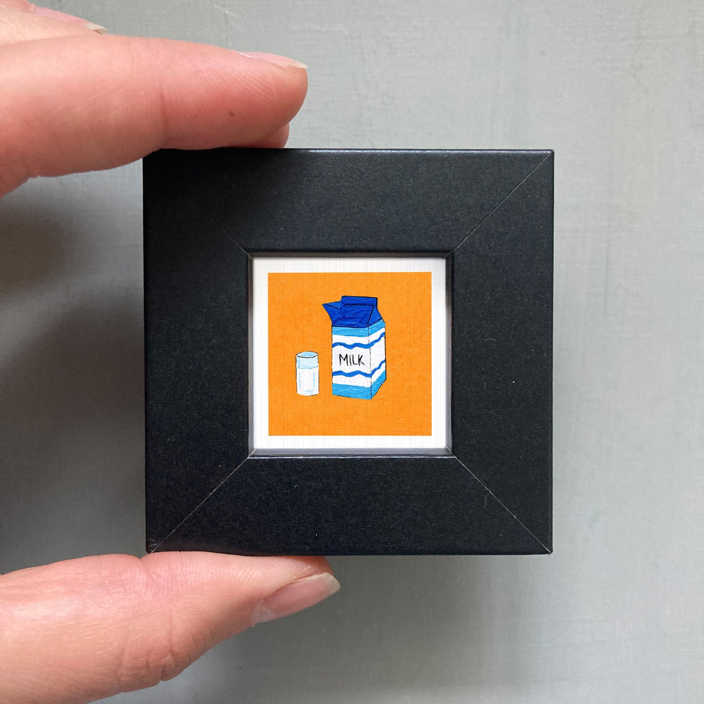 A hand is holding a small black square frame containing an illustration of a carton of milk and a small carton of milk.