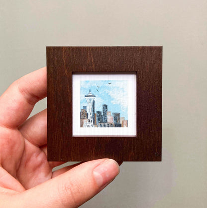 A hand is holding a small square picture frame containing a miniature painting of the Seattle skyline.