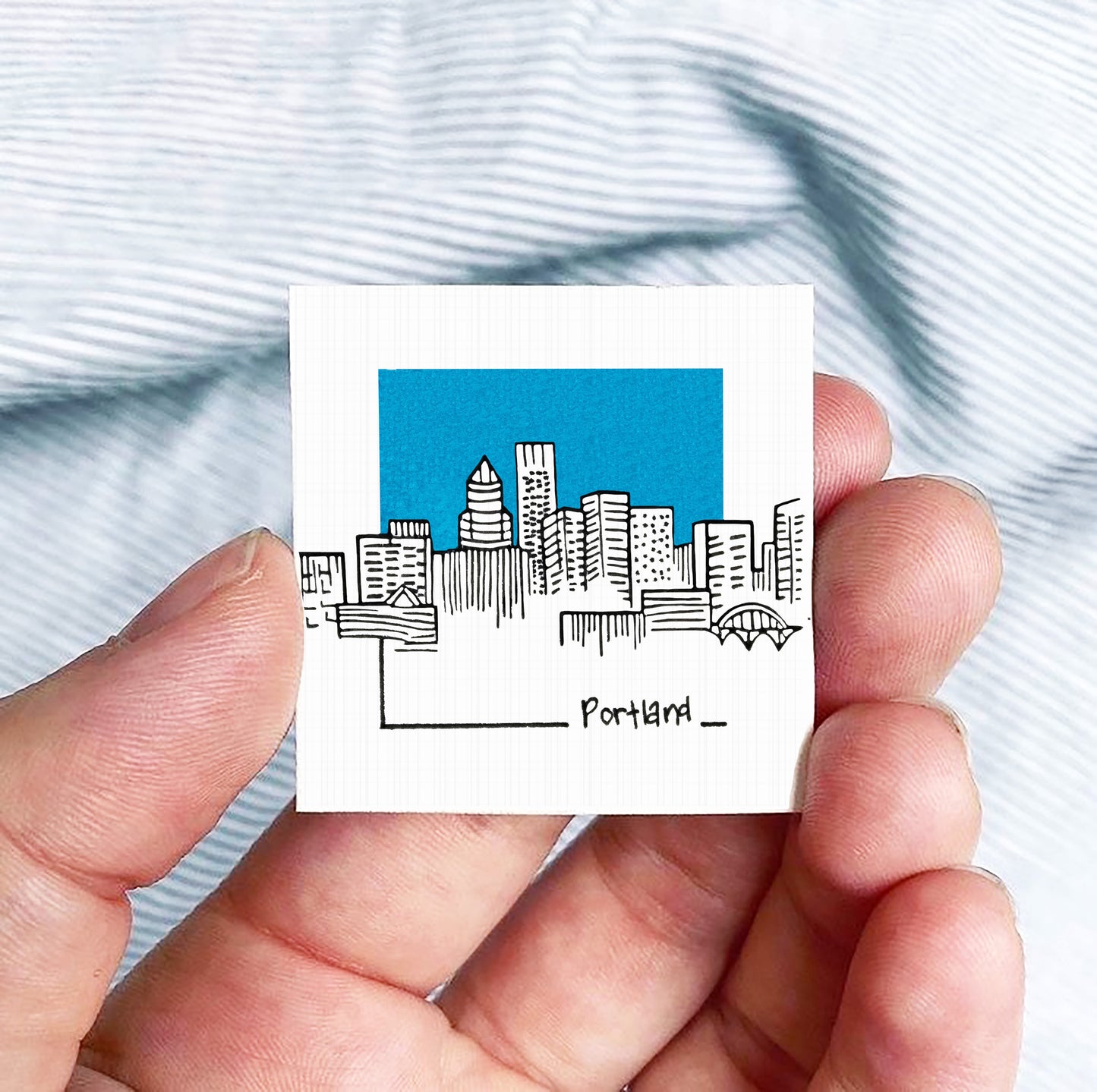 A hand holding a small square-shaped object with a drawing of a city skyline and the word "Portland" written on it.