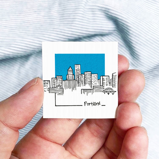 A hand holding a small square-shaped object with a drawing of a city skyline and the word "Portland" written on it.