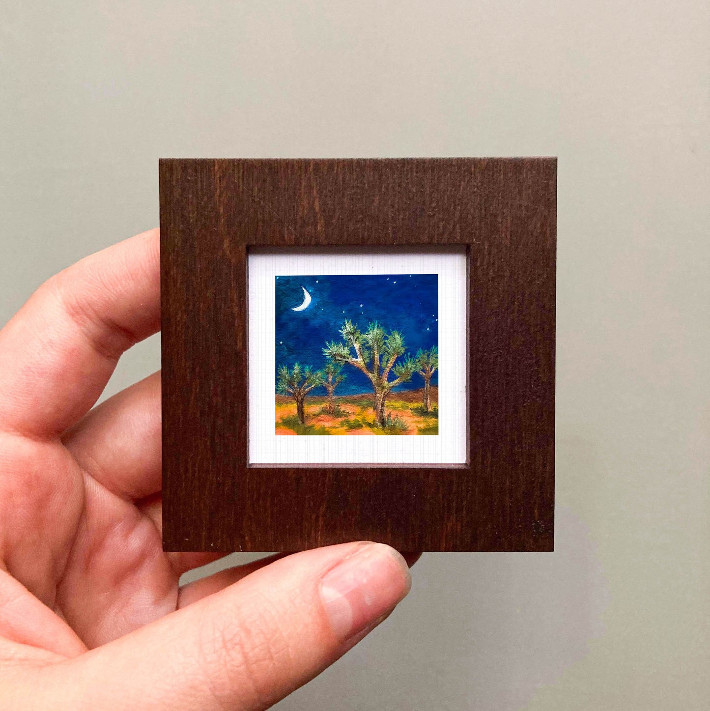 A hand is holding a small wooden frame containing a painting of a desert landscape with a crescent moon and stars in the night sky.