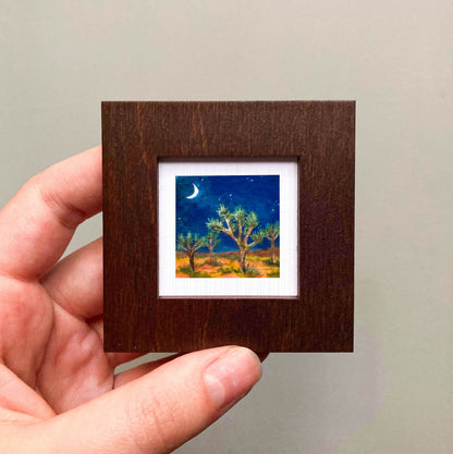 A hand is holding a small wooden frame containing a painting of a desert landscape with a crescent moon and stars in the night sky.