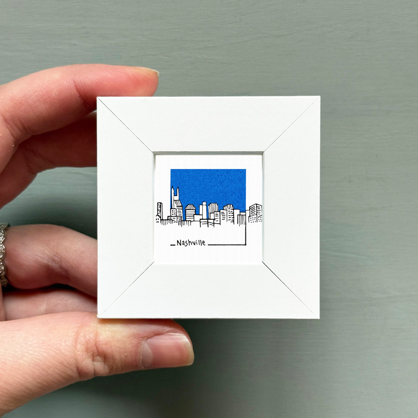 A hand is holding a small white square frame containing a blue illustration of a city skyline.