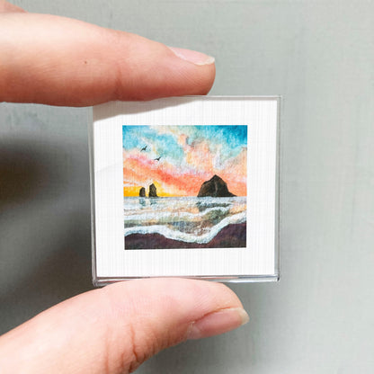 A person's hand holding a small square picture depicting a colorful sunset over a body of water.