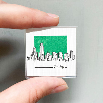 A hand holding a small square magnet with a drawing of a city skyline and the word "Chicago" written below it.