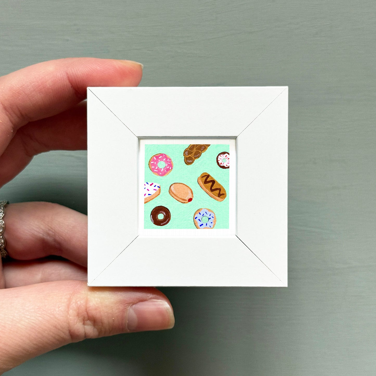 A hand is holding a small white square frame containing a collection of colorful donuts.