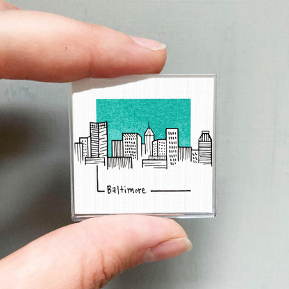 A hand holding a small square magnet with a drawing of a city skyline and the word "Baltimore" written below it.