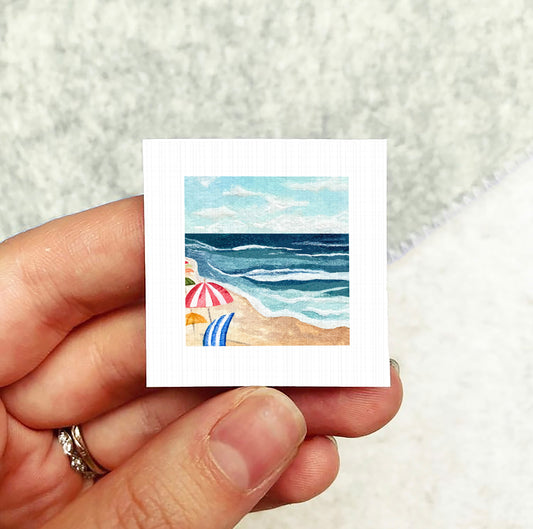 A hand holding a small square artwork depicting a beach scene with umbrellas and the ocean.