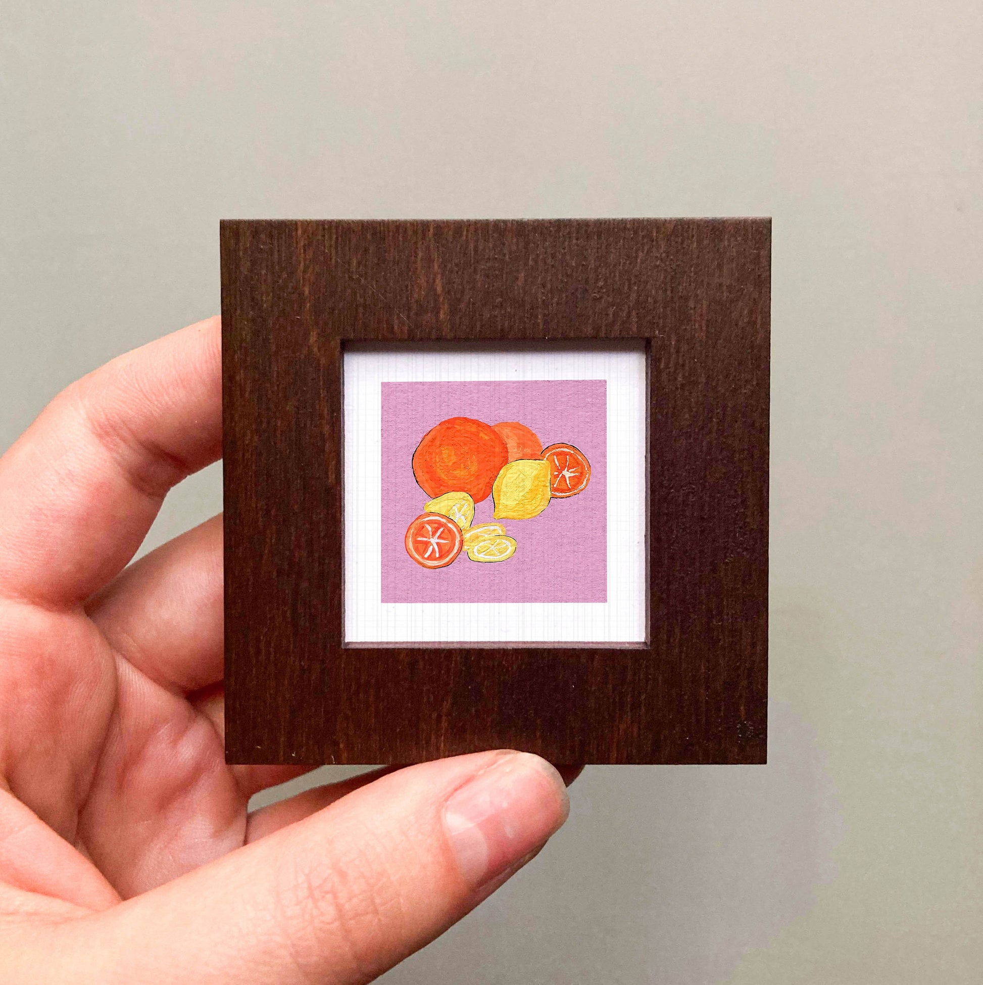 A hand is holding a small square frame containing an illustration of various fruits, including oranges and lemons.