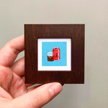 A hand is holding a small square frame containing an illustration of a can of soda and a glass of soda.