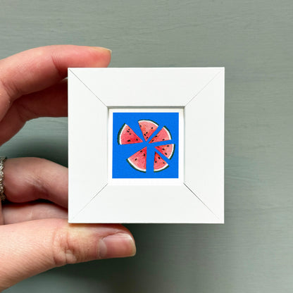 A hand is holding a small white square frame containing a vibrant illustration of watermelon slices.
