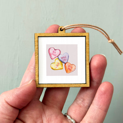 A hand holding a small wooden frame containing a picture of four heart-shaped candies with the text "Be Mine", "Miss You", "Love Me", and "Kiss Me" written on them.