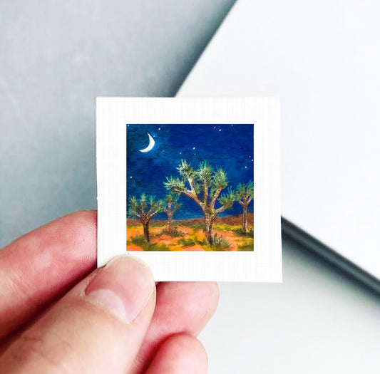 A hand holding a small square picture depicting a desert landscape with a crescent moon and stars in the night sky.