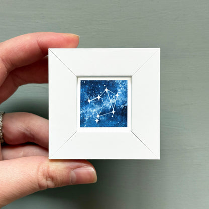 A hand is holding a small white frame containing a framed image of a starry night sky.