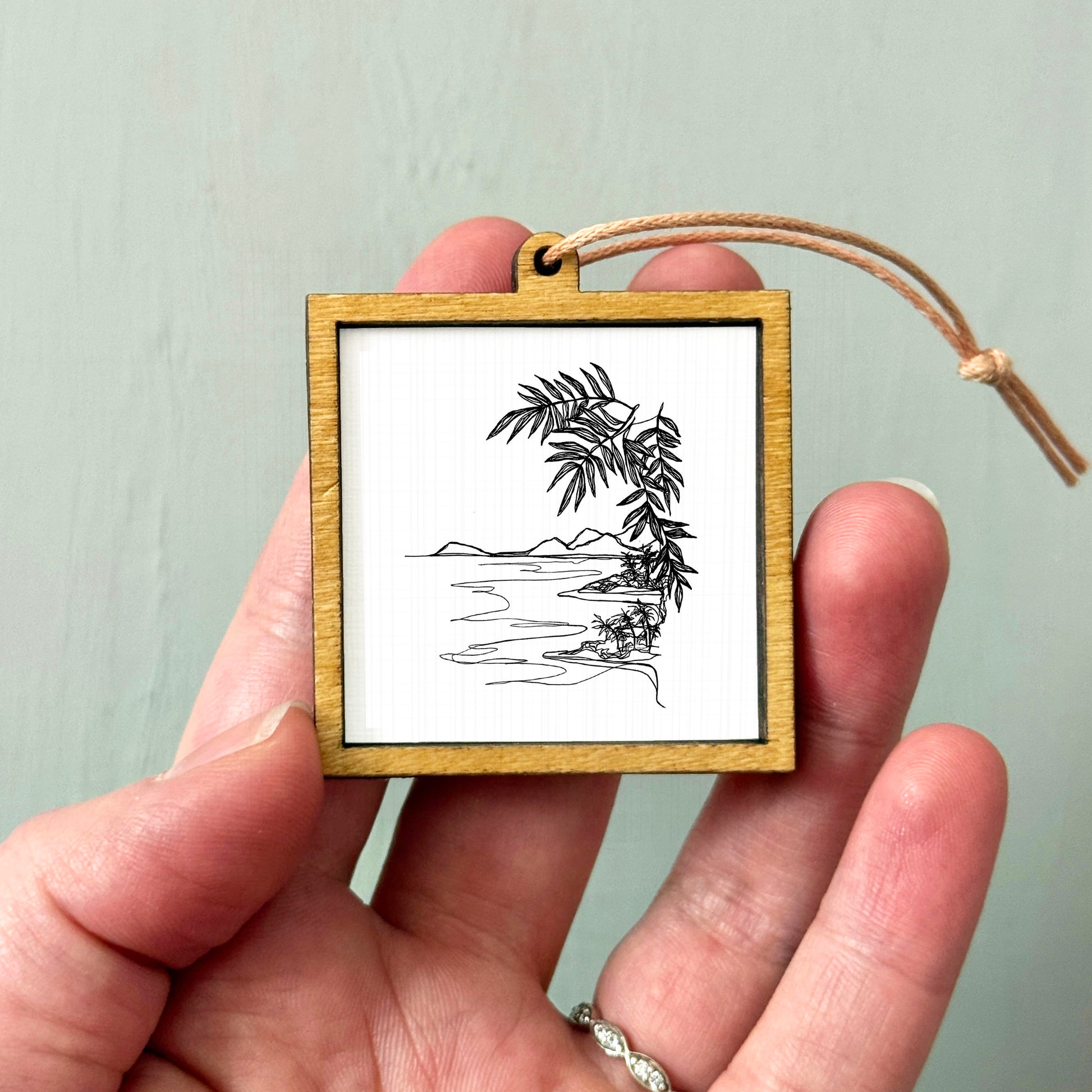 A hand holding a small wooden frame containing a black and white line drawing of a tropical landscape with palm trees and a body of water.