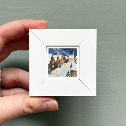 A hand is holding a small white square frame containing a painting of a snowy village scene.