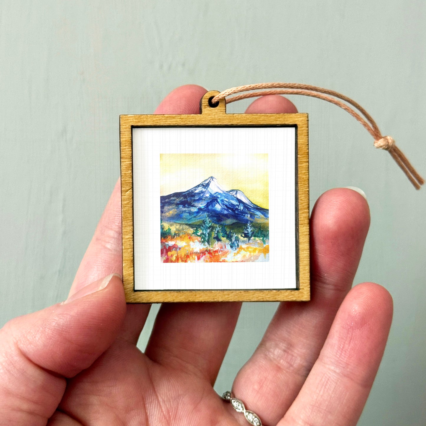A hand holding a small wooden frame containing a watercolor painting of a mountain landscape.
