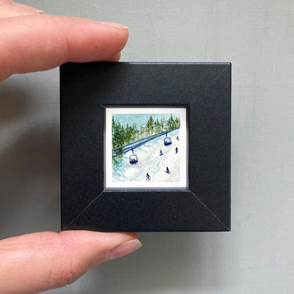 A person's hand is holding a small black frame containing a miniature painting of a ski lift and snowy landscape.