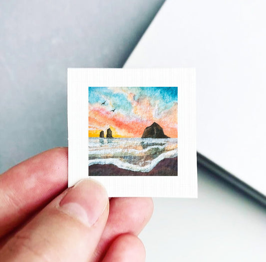 A hand is holding a small square artwork depicting a seascape with a rocky island in the background, set against a colorful sunset sky.