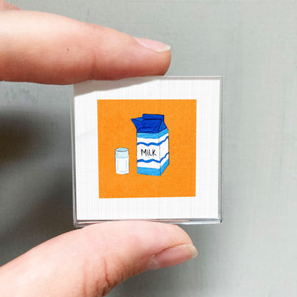 A person's hand holding a small square with an illustration of a carton of milk and a small container of milk.