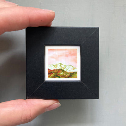 A person's hand is holding a small black frame containing a framed painting of a mountain landscape.