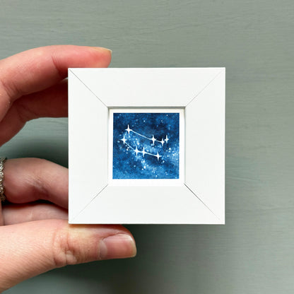 A hand is holding a small white square frame containing a watercolor painting of a starry night sky.