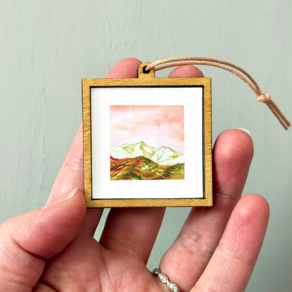 A hand holding a small wooden frame containing a framed landscape painting depicting mountains.