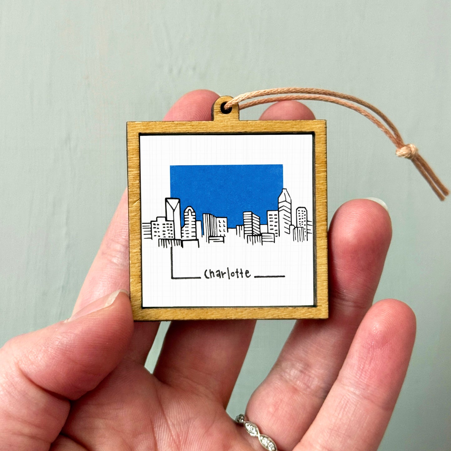 A hand holding a small wooden frame with a picture of a city skyline and the word "Charlotte" written on it.