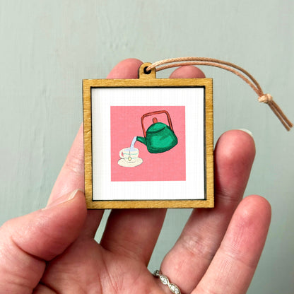 A person's hand holding a small wooden frame containing a framed illustration of a green teapot pouring tea.