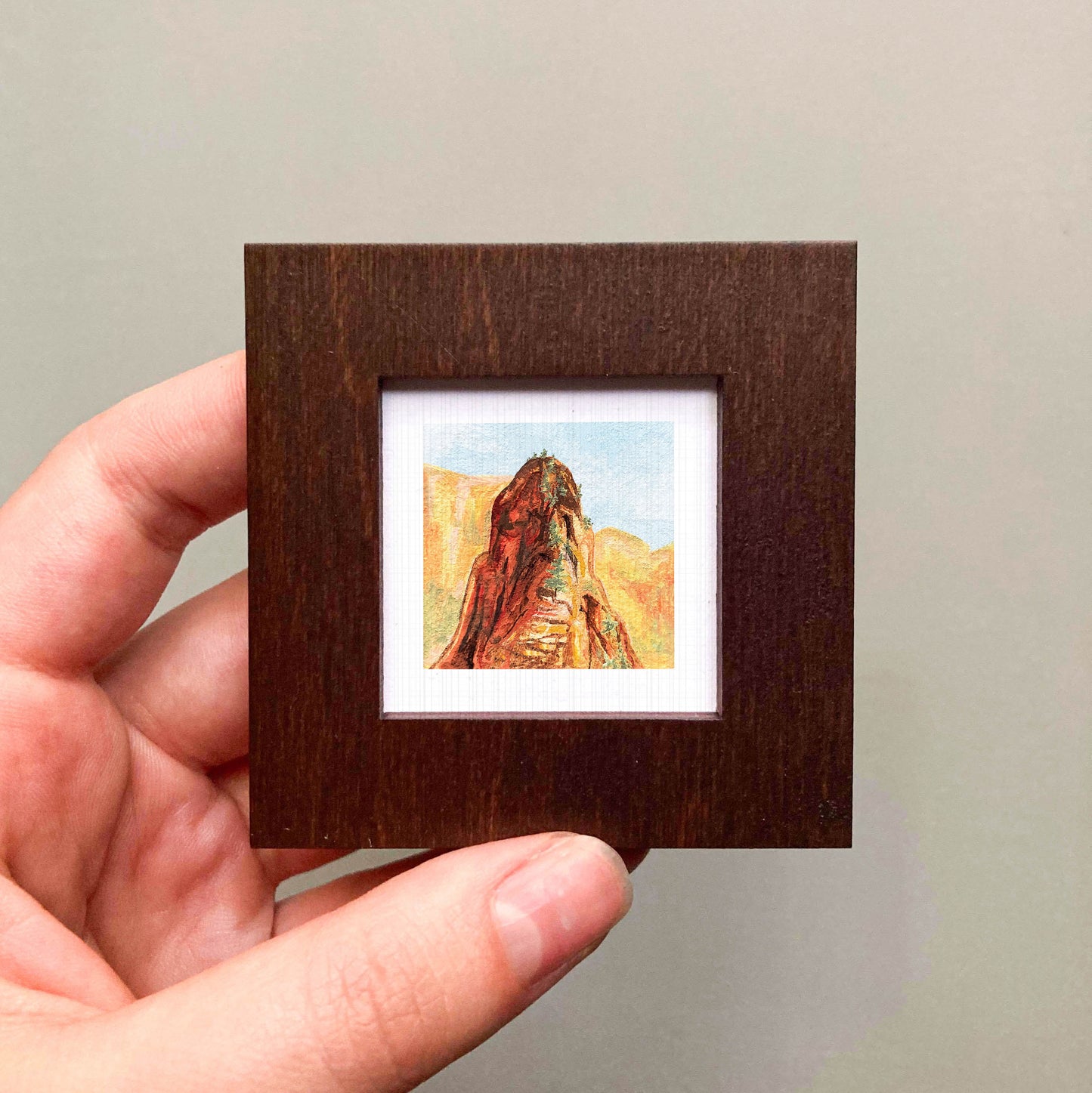 A hand is holding a small wooden frame containing a framed painting of a mountain landscape.