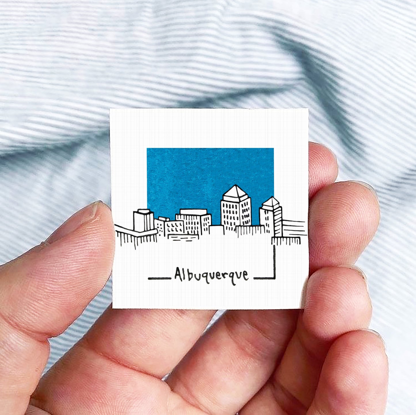 A hand holding a small square-shaped object with a drawing of a city skyline and the text "AlBUQUERQUE" written below it.