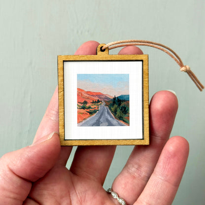 A hand holding a small wooden frame containing a painting of a winding road through a mountainous landscape.