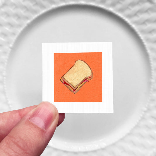 A hand is holding a small square with an illustration of a sandwich on it against a white background.